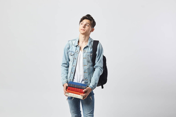 Young dark-haired guy with black backpack  on his shoulder dressed in a white t-shirt, jeans and a denim jacket holds books in his hands on the white background  in the studio - Photo, image