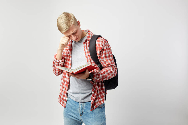 Sad blond guy with black backpack on his shoulder dressed in a white t-shirt, red checkered shirt and jeans reads a book on the white background  in the studio - Photo, Image