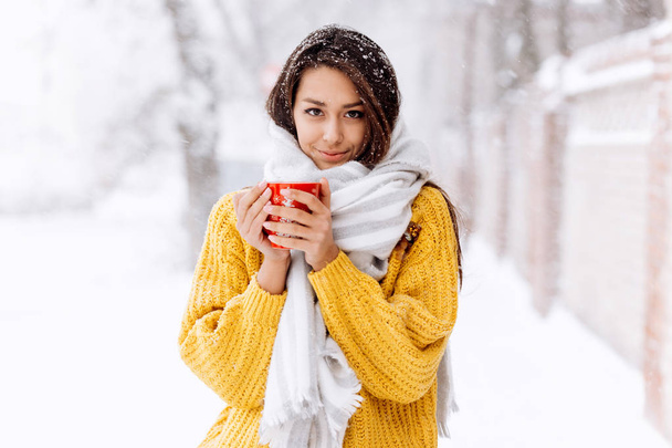 Cute dark-haired girl in a yellow sweater, jeans and a white scarf standing with a red mug on a snowy street on a winter day - 写真・画像