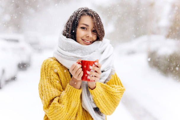Smiling dark-haired girl in a yellow sweater, jeans and a white scarf standing with a red mug on a snowy street on a winter day - Zdjęcie, obraz