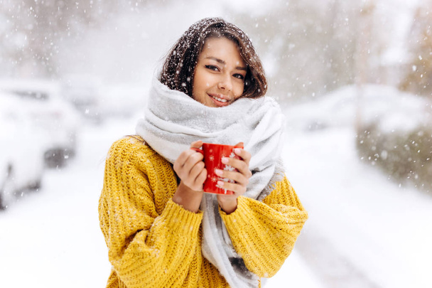 Smiling dark-haired girl in a yellow sweater, jeans and a white scarf standing with a red mug on a snowy street on a winter day - Foto, imagen
