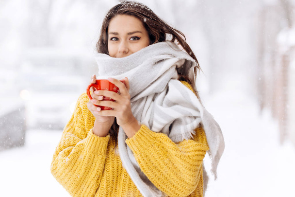 Nice dark-haired girl in a yellow sweater and a white scarf standing with a red mug on a snowy street on a winter day - Фото, изображение