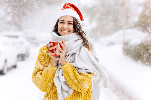 Smiling dark-haired girl in a yellow sweater, a white scarf  in Santa Claus hat is standing with a red mug on a snowy street on a winter day - Fotografie, Obrázek