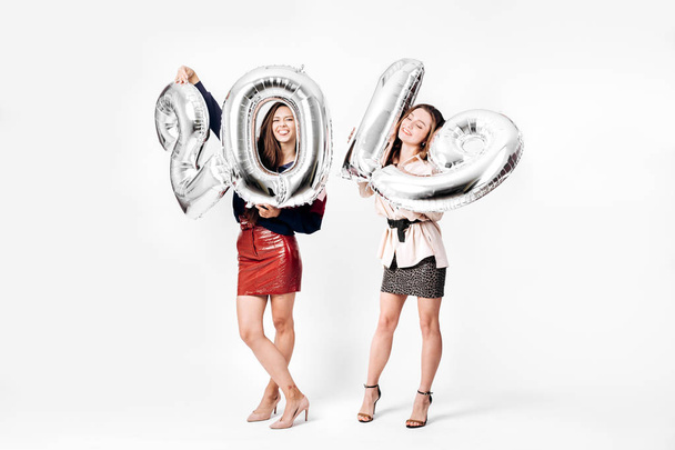 Two beautiful girls dressed in a stylish smart clothes are holding balloons in the shape of numbers 2019 on a white background in the studio - 写真・画像