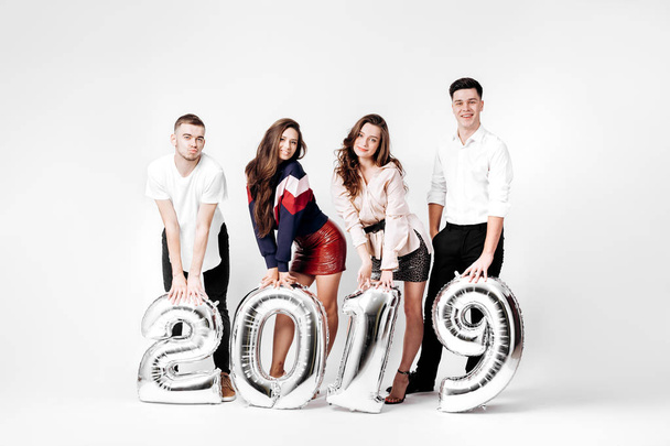 Group of happy friends of two girls and two guys dressed in stylish clothes are holding balloons in the shape of numbers 2019 on a white background in the studio - Fotoğraf, Görsel