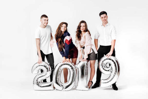Group of happy friends of two girls and two guys dressed in stylish clothes are holding balloons in the shape of numbers 2019 on a white background in the studio - Foto, Imagen