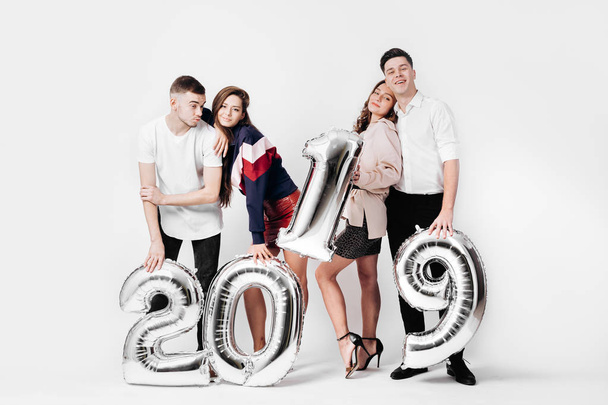 Grupo de amigos felices de dos chicas y dos chicos vestidos con ropa elegante están sosteniendo globos en forma de números 2019 sobre un fondo blanco en el estudio
 - Foto, Imagen