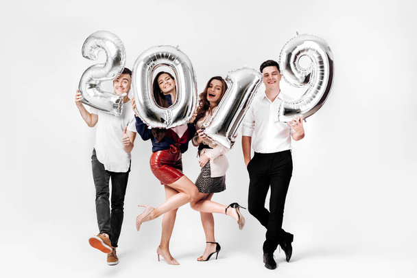 Merry friends two girls and two guys dressed in stylish clothes are holding balloons in the shape of numbers 2019 on a white background in the studio - Photo, Image