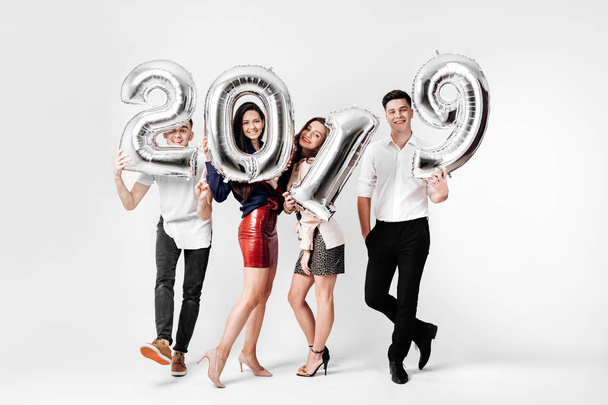 Merry friends two girls and two guys dressed in stylish clothes are holding balloons in the shape of numbers 2019 on a white background in the studio - Foto, Bild