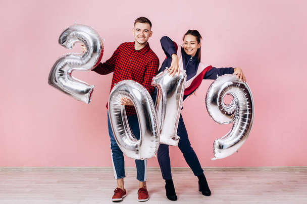 Funny friends girl and guy dressed in stylish clothes are holding balloons in the shape of numbers 2019 on a pink background in the studio - Фото, изображение