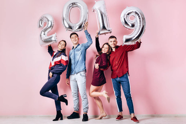 Happy company of two girls and two guys dressed in stylish clothes are holding balloons  in the shape of numbers 2019 over their heads on a pink background in the studio - Photo, Image