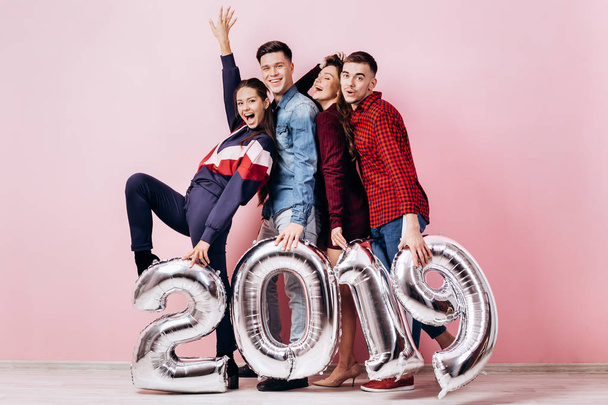 Happy company of two girls and two guys dressed in stylish clothes are holding balloons  in the shape of numbers 2019 on a pink background in the studio - Photo, Image