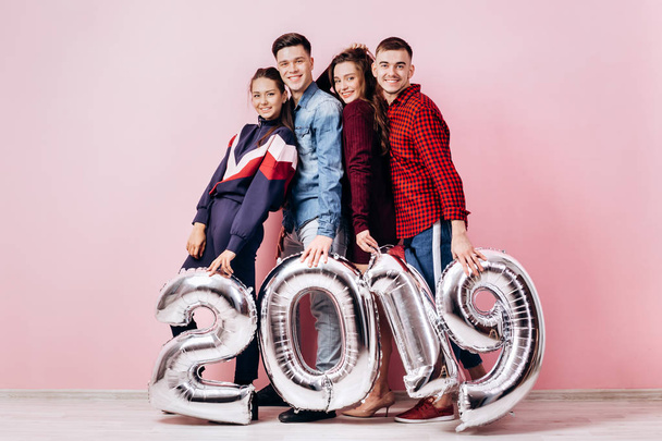 Happy company of two girls and two guys dressed in stylish clothes are holding balloons  in the shape of numbers 2019 on a pink background in the studio - Foto, Bild