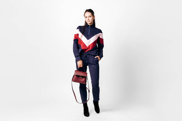 Girl dressed in sporty blue suit with a red and white print on a sweatshirt and heels poses with bag in her hands on the white background in the studio - Photo, Image