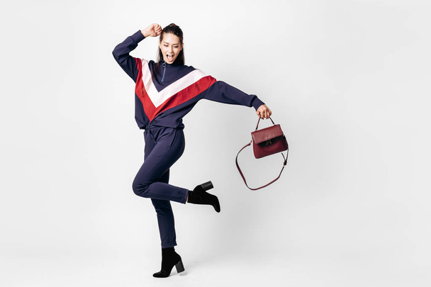 Funny girl dressed in sporty blue suit with a red and white print on a sweatshirt and heels poses with bag in her hands on the white background in the studio - Фото, изображение