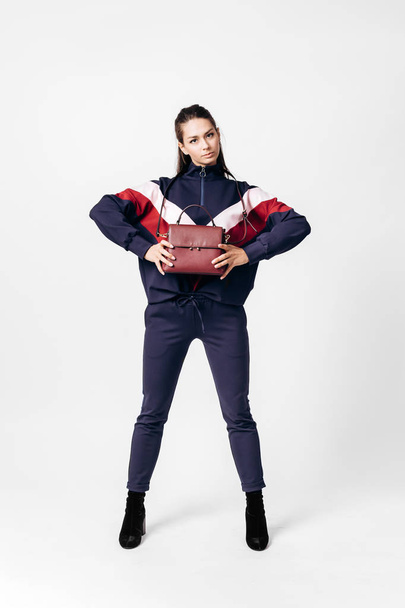 Girl dressed in sporty blue suit with a red and white print on a sweatshirt and heels poses with bag in her hands on the white background in the studio - Foto, afbeelding