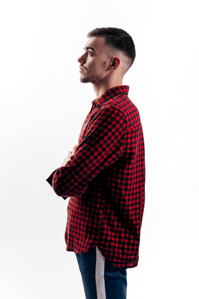 The guy dressed in red checkered shirt and jeans stands in profile on the white background in the studio - Foto, immagini