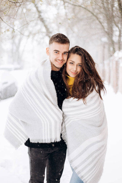 Charming dark-haired girl dressed in a yellow sweater and her boyfriend wrapped together in a gray scarf are standing in a snowy street on a winter day - Photo, Image