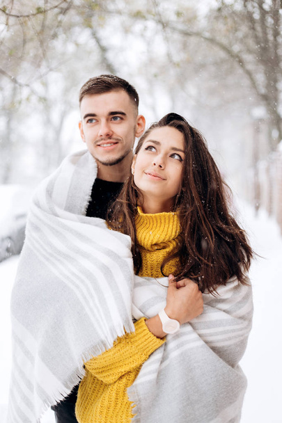 Charming dark-haired girl dressed in a yellow sweater and her boyfriend wrapped together in a gray scarf are standing in a snowy street on a winter day - Φωτογραφία, εικόνα