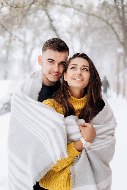 Charming dark-haired girl dressed in a yellow sweater and her boyfriend wrapped together in a gray scarf are standing in a snowy street on a winter day - Zdjęcie, obraz