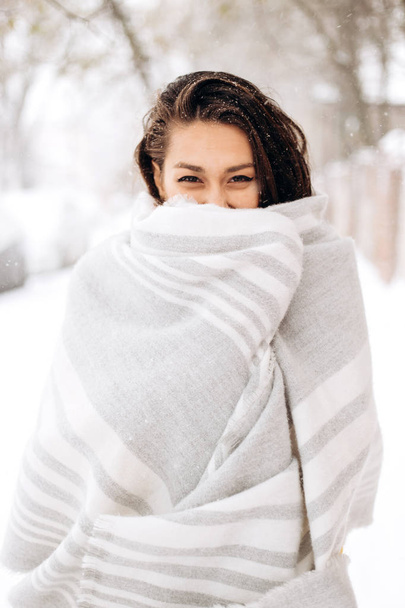 Charming dark-haired girl wrapped in a gray scarf is standing in a snowy street on a winter day - Zdjęcie, obraz