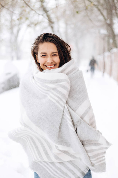 Smiling dark-haired girl wrapped in a gray scarf is standing in a snowy street on a winter day - Zdjęcie, obraz