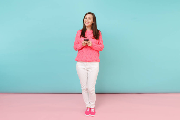 Full length portrait of woman in knitted sweater, white pants texting sms on cellphone isolated on bright pink blue pastel wall background in studio. Fashion lifestyle concept. Mock up copy space - Photo, Image