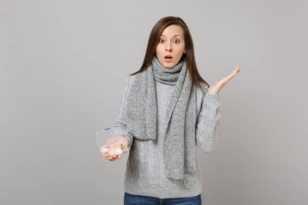 Shocked young woman in gray sweater, scarf spreading hands, holding daily pill box isolated on grey background. Healthy lifestyle, ill sick disease treatment, cold season concept. Mock up copy space - Photo, Image