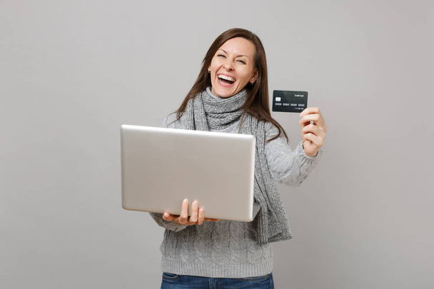Смеющаяся молодая женщина в сером свитере, шарф, работающий на компьютере ноутбука, держащий кредитную карту, изолированную на сером фоне стены. Консультации по лечению здорового образа жизни, концепция холодного сезона
 - Фото, изображение