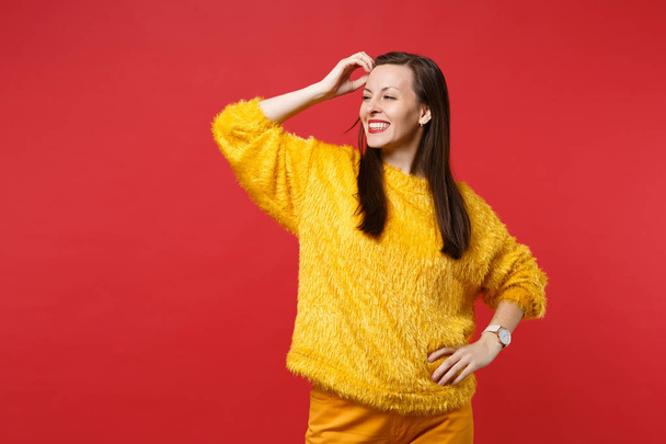 Радостная молодая женщина в желтом меховом свитере смотрит в сторону, стоя с рукой возле лба, изолированного на ярко-красном фоне стены в студии. Люди искренние эмоции концепции образа жизни. Пространство для копирования
 - Фото, изображение