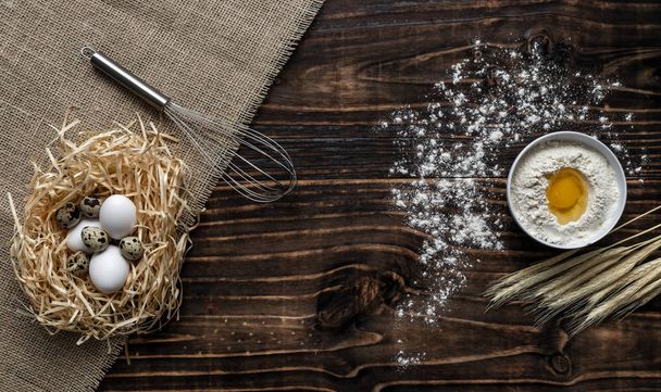 Мука в белой миске, темный деревянный фон с колосьями пшеницы, яйца в соломенном гнезде, вид сверху с копировальным пространством. ингредиенты для хлебобулочных изделий
 - Фото, изображение