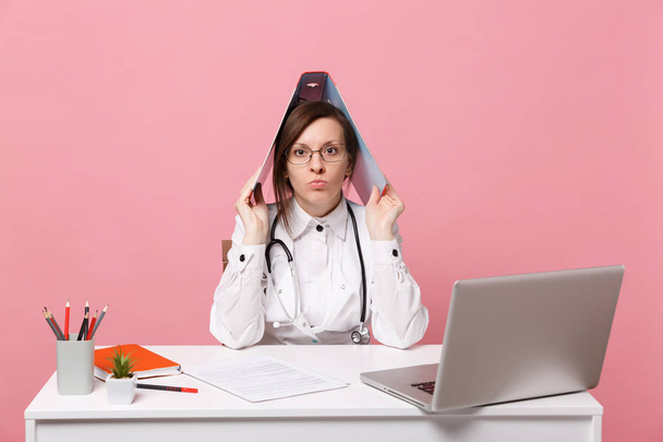 Female doctor sit at desk work on computer with medical document hold folder in hospital isolated on pastel pink background. Woman in medical gown glasses stethoscope. Healthcare medicine concept - Fotografie, Obrázek