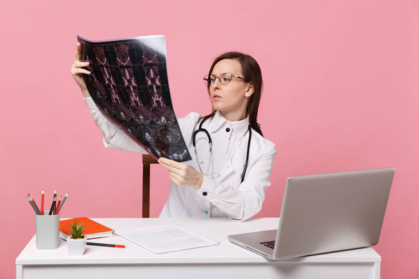 Female doctor sit at desk work on computer with medical document hold x-ray in hospital isolated on pastel pink wall background. Woman in medical gown glasses stethoscope. Healthcare medicine concept - Photo, Image