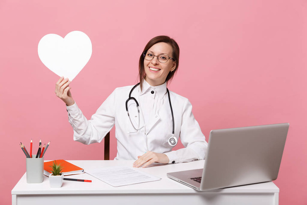 Lékařka si sedne na stole práci na počítači s lékařských dokumentu podržte srdce v nemocnici izolován na pastelově růžové zdi pozadí. Žena v lékařské šaty brýle stetoskop. Koncepce zdravotnictví medicína - Fotografie, Obrázek