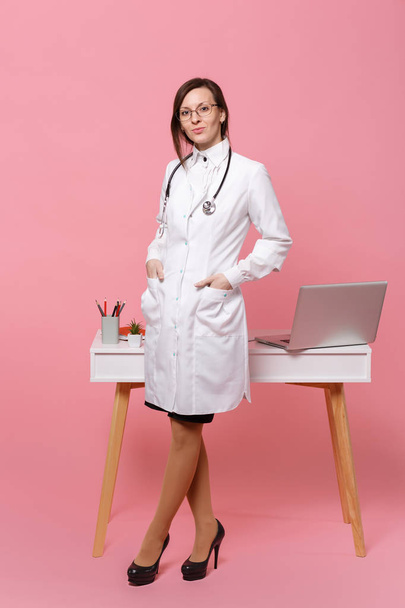 Ładna kobieta lekarz podstawka z przodu biurko z komputerem pc, dokument medyczny w szpitalu na białym tle na tle pastelowy róż. Kobieta w sukni medyczne okulary stetoskop. Opieki zdrowotnej medycyny pojęcie - Zdjęcie, obraz