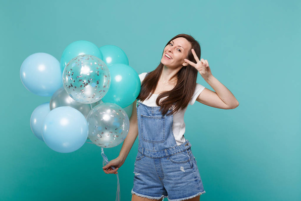 Smiling young woman in denim clothes showing victory sign, celebrating and holding colorful air balloons isolated on blue turquoise wall background. Birthday holiday party, people emotions concept - Photo, image