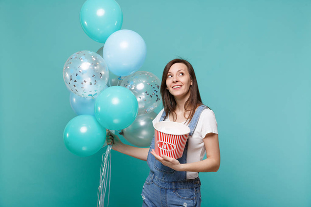 Pensive young woman in denim clothes looking up holding bucket of popcorn celebrating with colorful air balloons isolated on blue turquoise background. Birthday holiday party, people emotions concept - Photo, image