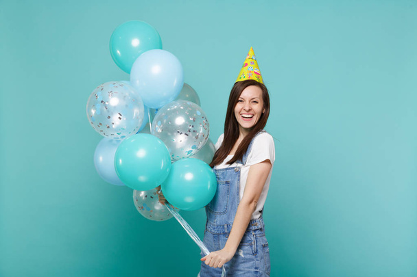 Portrait of cheerful young woman in denim clothes, birthday hat celebrating, holding colorful air balloons isolated on blue turquoise wall background. Birthday holiday party, people emotions concept - Photo, Image