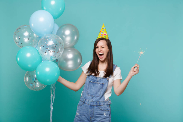 Fröhlich blinkende junge Frau mit Geburtstagsmütze und brennender Wunderkerze, die mit bunten Luftballons auf türkisblauem Hintergrund feiert. Geburtstagsparty, Menschen Emotionen Konzept - Foto, Bild