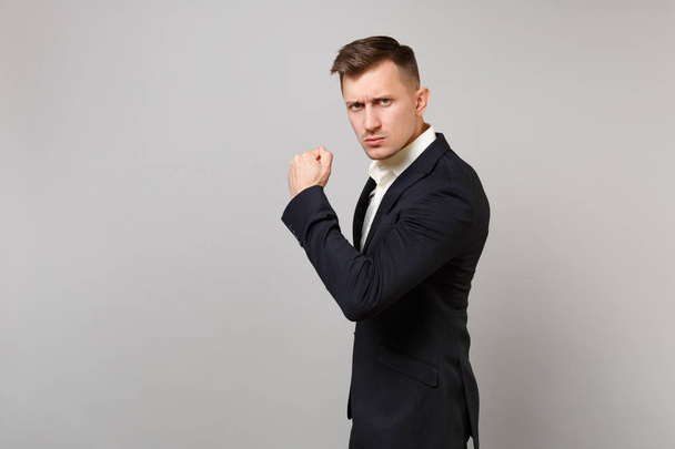 Strong young business man in classic black suit and shirt clenching fist, showing biceps muscles isolated on grey background in studio. Achievement career wealth business concept. Mock up copy space - Foto, Imagen