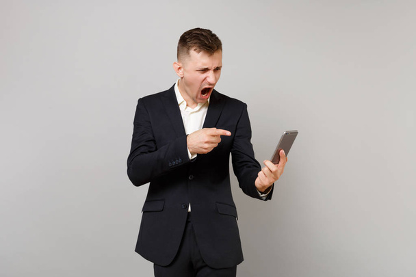 Angry business man swearing screaming, pointing index finger on on mobile phone in hand, making video call isolated on grey background. Achievement career wealth business concept. Mock up copy space - Photo, Image