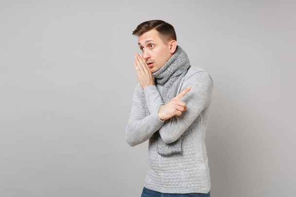 Young man in gray sweater, scarf whispers gossip, tells secret with hand gesture, point index finger aside isolated on grey background. Healthy lifestyle, people sincere emotions, cold season concept - Photo, Image