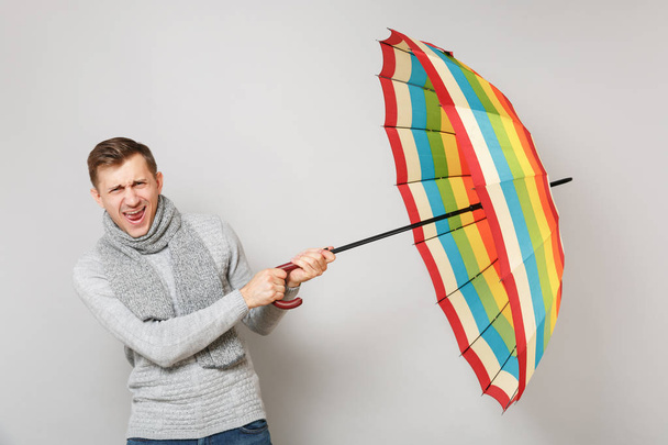 Χαρούμενος νεαρός άνδρας σε γκρι πουλόβερ, κασκόλ ουρλιάζοντας, κρατώντας ομπρέλα που απομονώνονται σε γκρι φόντο. Υγιή τρόπο ζωής, άνθρωποι ειλικρινή συναισθήματα, κρύο σεζόν έννοια. Κοροϊδεύετε αντίγραφο χώρου - Φωτογραφία, εικόνα