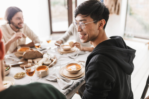 Junger fröhlicher asiatischer Mann mit dunklen Haaren, Brille und Kapuzenpulli sitzt am Tisch und schaut fröhlich zur Seite. Gruppe internationaler Freunde beim gemeinsamen Mittagessen im Café - Foto, Bild