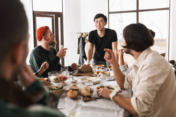 若いメガネ、喜んでカメラの検索テーブルに寄りかかって黒 t シャツのアジア人男性の笑顔します。居心地の良いカフェでランチの時間を一緒に過ごす魅力的な国際的な友達のグループ - 写真・画像