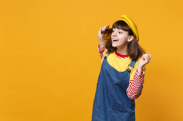 Nieuwsgierig meisje tiener in Franse baret, denim sundress opzij kijken aandachtig ver afstand afgezonderd op gele muur achtergrond. Mensen oprechte emoties, lifestyle concept. Bespotten kopie databaseruimte - Foto, afbeelding