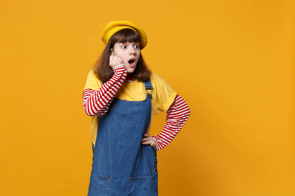Amazed girl teenager in french beret looking aside, keeping mouth wide open, putting hands on cheeks isolated on yellow wall background. People sincere emotions lifestyle concept. Mock up copy space - Photo, Image