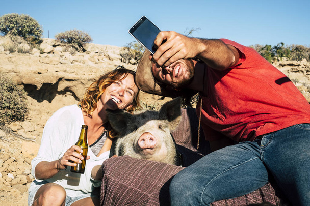 Alternative family or friends with young man and woman smiling while take a selfie with a domestic pig in the middle - everybody smile for the camera - sunny day and unusual use of phone camera technology - funny animal - Photo, Image