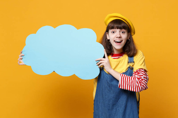 Excited girl teenager in french beret, denim sundress hold blue empty blank Say cloud, speech bubble isolated on yellow wall background. People sincere emotions, lifestyle concept. Mock up copy space - Photo, Image