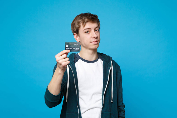 Portrét pohledný mladý muž v neformálním oblečení drží bankovní kartou izolované na pozadí modré zdi ve studiu. Lidé upřímné emoce, stylu života. Vysmívat se kopie prostor. Reklamní plocha - Fotografie, Obrázek